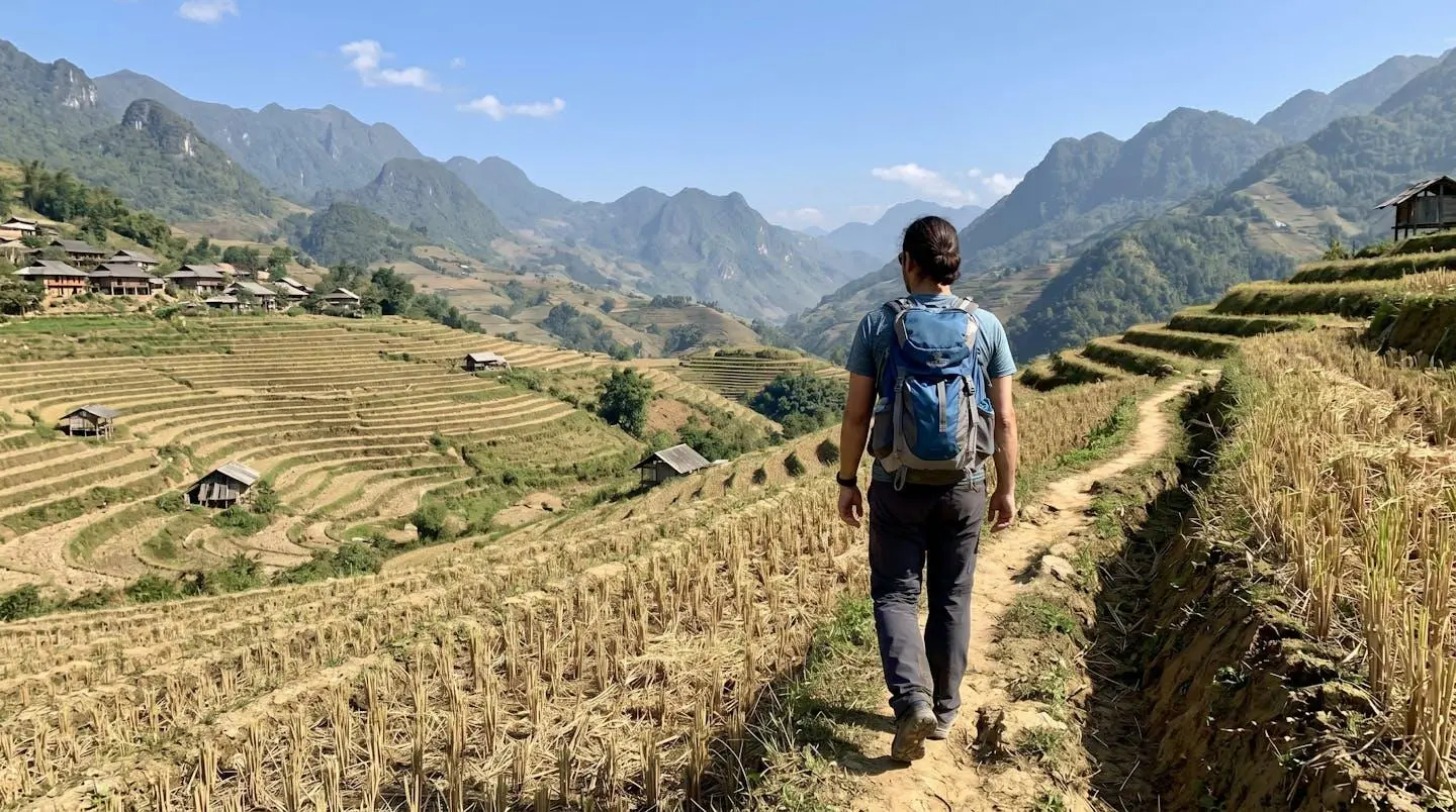 Femme de dos avec sac à dos de randonnée marchant sur un sentier étroit bordant des rizières en terrasses, montagnes brumeuses à l'horizon